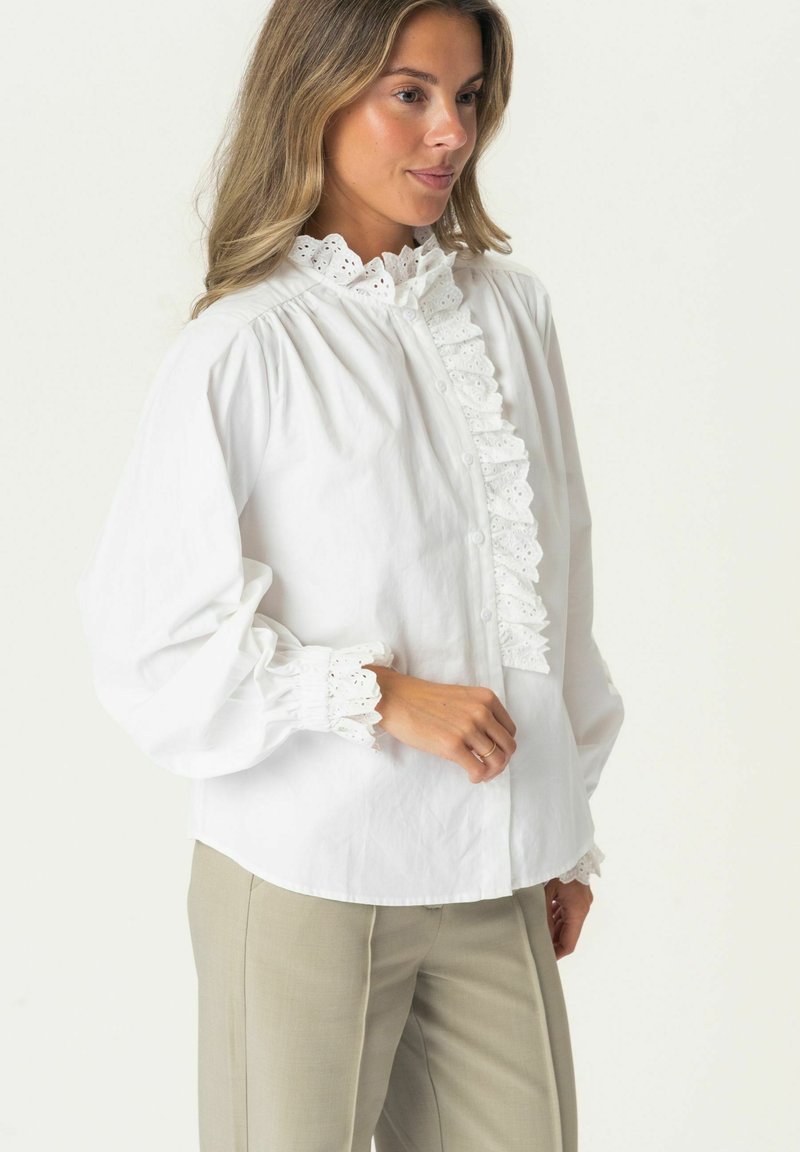 White cotton blouse with a high ruffled collar, front button closure, and lace trim on cuffs. Long sleeves and soft, smooth texture.