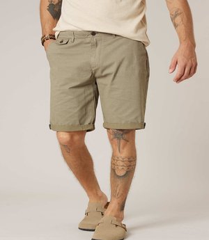Man die beige gebloemde korte broek, beige instapschoenen en een lichtbeige shirt draagt, met tatoeages op armen en benen zichtbaar.
