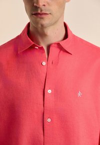 Camisa de lino color coral con cuello abotonado, que presenta botones blancos y un pequeño logo bordado en el lado izquierdo del pecho.