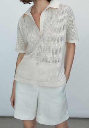 Persoon die een beige, kortmouwig polohemd van mesh breisel en losse witte korte broek draagt, staande tegen een lichtgrijze achtergrond.