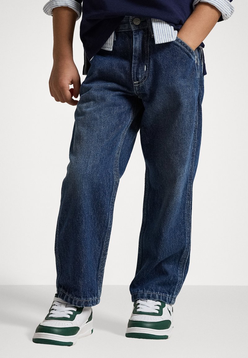 Calças de ganga azul-escuro com um ajuste largo e textura desgastada, combinadas com ténis brancos com detalhes em verde.