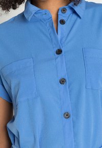 Chemise bleue à manches courtes en tissu lisse, avec col, deux poches poitrine et ourlet arrondi. Des boutons noirs soulignent le design.