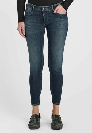 Person trägt dunkelblaue Skinny Jeans und schwarze Slipper, mit einer Hand in der Tasche und einem schwarzen Langarmoberteil.