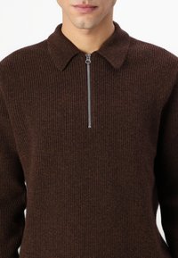Pull en maille côtelée marron avec un col montant et une fermeture éclair à l'avant. Le matériau semble doux avec une finition texturée et des variations subtiles de couleur.