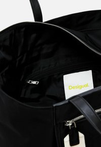 Black tote bag with a zippered opening, featuring a smooth exterior, leather handles, and an interior zip pocket. Visible brand tag in yellow.
