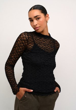 Black lace long-sleeve top featuring a layered design with a high neckline and scalloped edges. Worn over a fitted camisole.
