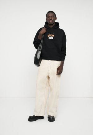 Felpa nera con stampa grafica, pantaloni beige a gamba larga e scarpe nere con suola spessa. Porta una borsa a tracolla in pelle nera.