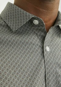 Une chemise à col dans des tons atténués présente un motif géométrique en losanges. Le tissu est lisse avec des fermetures à boutons et un design de col structuré.