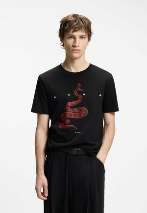 Junger Mann trägt ein schwarzes T-Shirt mit einem roten, gewundenen Schlangengrafik und dem Schriftzug „HUGO“ auf der Brust, steht mit einer Hand in der Tasche.