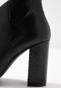 Schwarze Ankle Boots mit Absatz, die ein strukturiertes Schlangenlederdesign aufweisen, sowie einen soliden, hohen Blockabsatz und ein glattes, elegantes Obermaterial.