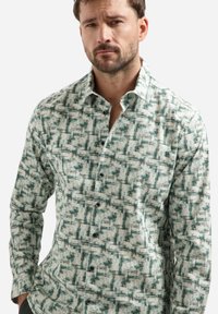 Man in een overhemd met lange mouwen en een groen-wit abstract vierkant patroon, staande met handen in de zakken.