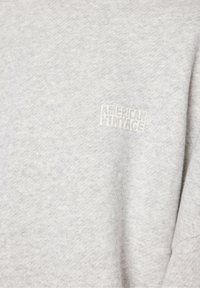 Sivá kapuca iz bazi z mehkim, puhastim materialom. Vsebuje mali bel logo "AMERICAN VINTAGE" na zgornjem levem predelu prsi.