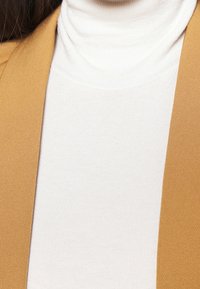 Bílý rolák vyrobený z žebrované látky, spárovaný s jednobarevným hnědým blazerem. Detailní pohled zaměřující se na strukturu a vrstvení.