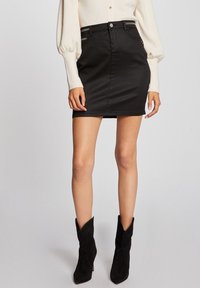 Minifalda de mezclilla negra con corte ajustado, con bolsillos frontales y detalles plateados. Combinada con una blusa blanca de manga larga y botas negras de tacón.
