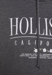 Grå hoodie med "HOLLISTER CALIFORNIA" i vita bokstäver, med blommiga accenter och namn på kaliforniska städer listade nedan. Mjuk textur.