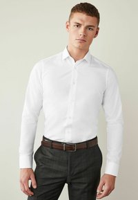 Cămașă albă cu mâneci lungi, cu guler clasic, asociată cu pantaloni cu carouri închise și o curea din piele maro cu prindere argintie.