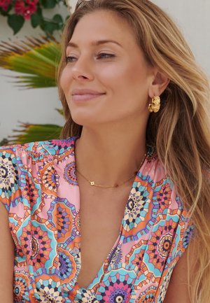 Smiling woman with long blonde hair wears colorful patterned top, gold hoop earrings, and a delicate gold necklace, with green foliage in background.