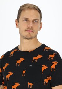 Musta T-paita, jossa on toistuva oranssi hirvipaino ja rintatasku, jossa on etiketti. Paita on valmistettu pehmeästä ja hengittävästä kankaasta.