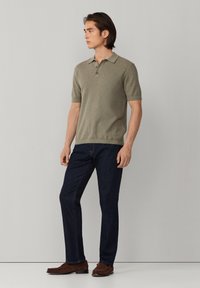 Muškarac obučen u olivno zelenu polo majicu kratkih rukava, tamnoplave traperice i smeđe mokasine, stoji naslonjen na svijetlosivu zidnu površinu, gleda lijevo.