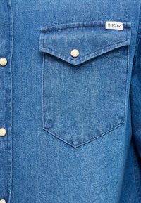 Helevalge teksasärk, millel on terava nurgaga taskuklapid, trukknööbi kinnis ja õmmeldud "Mustangi" silt õmblusel õlal.