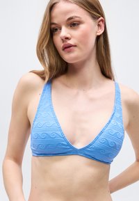 Νέα γυναίκα που φοράει ένα ανοιχτό μπλε μπικίνι με σχέδια κυμάτων, κοιτάζοντας ελαφρώς προς τα κάτω και πλάγια σε ένα απλό φόντο.