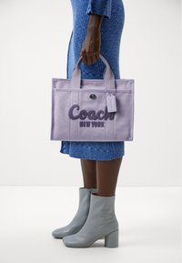 Levendula színű vászontáska "Coach New York" sötétlila hímzésével, dupla fülekkel és függő címkével. Texturált felület és formázott alak.