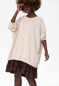 Beżowy, oversize sweter z miękkiego, teksturowanego materiału, z rękawami w kształcie nietoperza, zestawiony z ciemnoczerwonym, warstwowym będącym spódnicą z koronkowym wykończeniem.