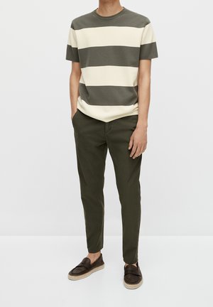 Homme portant une chemise à manches courtes rayée verte et blanche, un pantalon vert olive et des chaussures marron foncé sans lacets, debout les mains dans les poches.