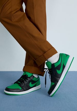 Osoba w brązowych spodniach i białych skarpetkach z czarno-zielonymi butami sportowymi Nike stojąca na niebieskiej podłodze.