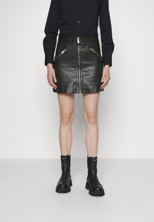 Femme portant une mini-jupe en cuir noir avec fermeture éclair à l'avant et poches zippées, associée à des bottines noires et une chemise noire boutonnée.