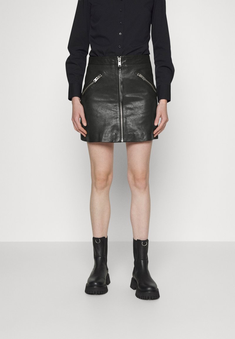 Femme portant une mini-jupe en cuir noir avec fermeture éclair à l'avant et poches zippées, associée à des bottines noires et une chemise noire boutonnée.