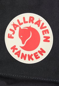 Piros kör alakú folt egy stilizált róka ikonként és a "FJÄLLRÄVEN KÅNKEN" felirattal, fekete szövet háttérrel.