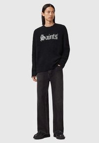 Czarny dzianinowy sweter z napisem "Saints" w białych literach, zestawiony z luźnymi czarnymi jeansami i czarnymi butami. Gładka tekstura, swobodny krój.