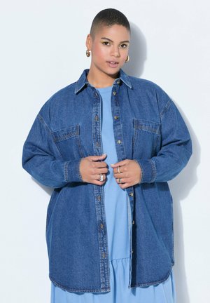 Osoba s krátkými vlasy, nosící zlaté kruhové náušnice a prsteny, oblečená v modrých šatech a nadměrné džínové bundě, pózující před bílou zdí.