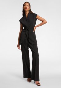 Czarny kombinezon z talią w paski, krótkimi rękawami i szerokimi nogawkami. Gładka tkanina z delikatnym drapowaniem i dwoma przednimi kieszeniami.