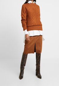 Hrubý svetr na pletení v rezavé barvě s copánkovým vzorem, vrstvený přes bílou košili. Hnědá semišová sukně s předním rozparkem, doplněná vzorovanými hnědými botami.