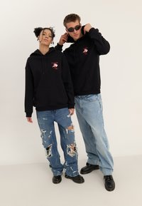 Two people wearing black hoodies with red floral designs; one adjusts hoodie, both wear ripped and loose jeans with black shoes.