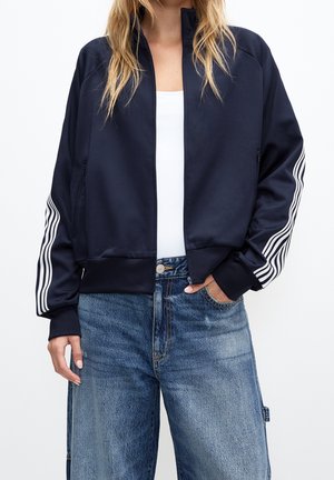 Kvinde iført en mørkeblå lynlåsjakke med hvide striber på ærmerne, hvid top og blå denimjeans med den ene hånd i lommen.