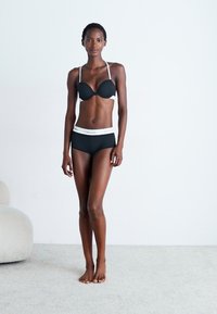Svart bh med formerte kopper og tynne hvite stropper, parret med svarte boy shorts med hvit midjebånd. Modellen står på et nøytralt teppe.