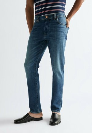 Mężczyzna w średnio niebieskich dżinsach, czarnych butach wsuwanych i granatowo-różowej koszulce bez rękawów, stojący z jedną ręką w kieszeni.