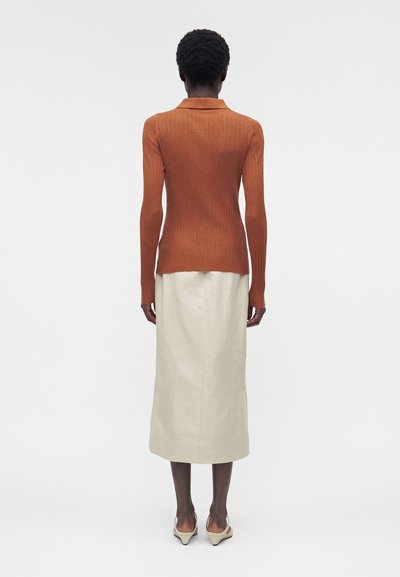Άτομο που φαίνεται από πίσω, φοράει εφαρμοστή μακρυμάνικη μπλούζα σε σκουριά απόχρωση, κρεμ midi φούστα και μπεζ παπούτσια με χαμηλό τακούνι σε λευκό φόντο.
