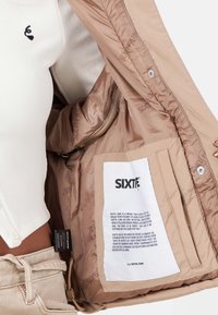 Beige isolierte Jacke mit weichem Innenfutter, ausgestattet mit einem Etikett mit Markeninformation, einer gefütterten Tasche und subtiler Branding-Optik.