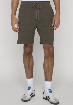Mann trägt olivgrüne Waffelstrick-Shorts mit Kordelzug, weiße Sportsocken und weiße Sneaker mit schwarzen Streifen und blauen Logopatches.