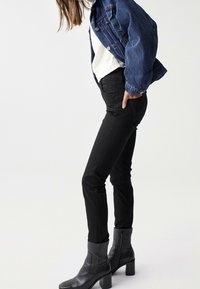 Kvinna som bär svarta slim fit-byxor, svarta klackade ankelstövlar, vit topp och en kort blå denimjacka mot en enfärgad bakgrund.