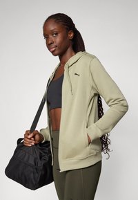 Olivgrön ziphoodie med mjuk textur, med en svart logotyp på bröstet. Modellen håller en svart väska med nätdetaljer.