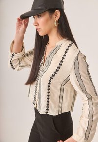 Blusa beige recortada con bordado geométrico negro, mangas largas, escote en forma de V y cierre de botones, combinada con pantalones negros de cintura alta.