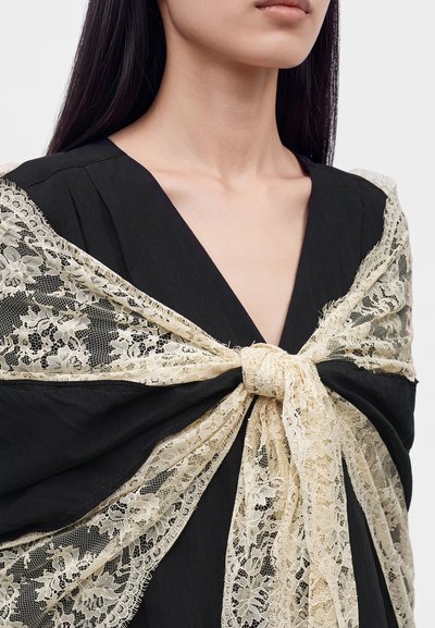 Femme portant une robe noire à col en V avec un châle en dentelle crème noué en un nœud autour des épaules sur un fond clair.