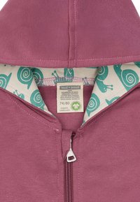 Felpa con zip color bordò, dal tessuto morbido; all'interno presenta una fodera crema con un motivo di lumache verdi. L'etichetta mostra informazioni sul cotone biologico.