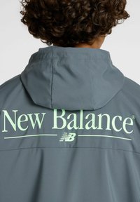 Persona con cabello rizado que lleva una chaqueta con capucha gris con un gran logo de "New Balance" en verde claro en la parte posterior.