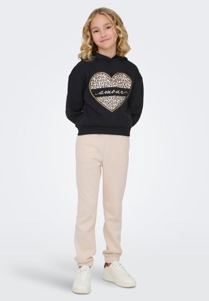 Jeune fille aux cheveux blonds bouclés portant un sweat à capuche noir avec un cœur imprimé léopard et le texte « amour », un pantalon beige et des baskets blanches.